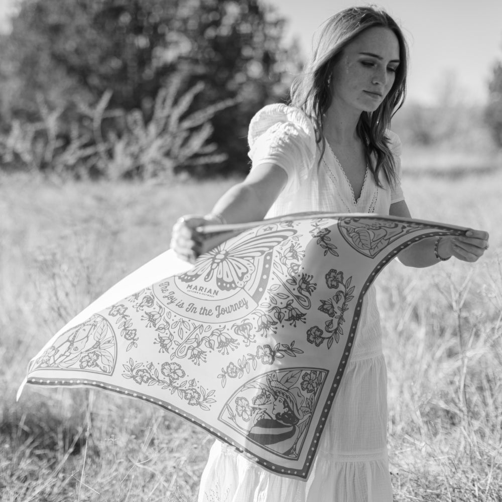 A woman in a white dress spreading her arms gracefully amidst a blooming field, wearing the Butterfly Scarf by Shop Marian.