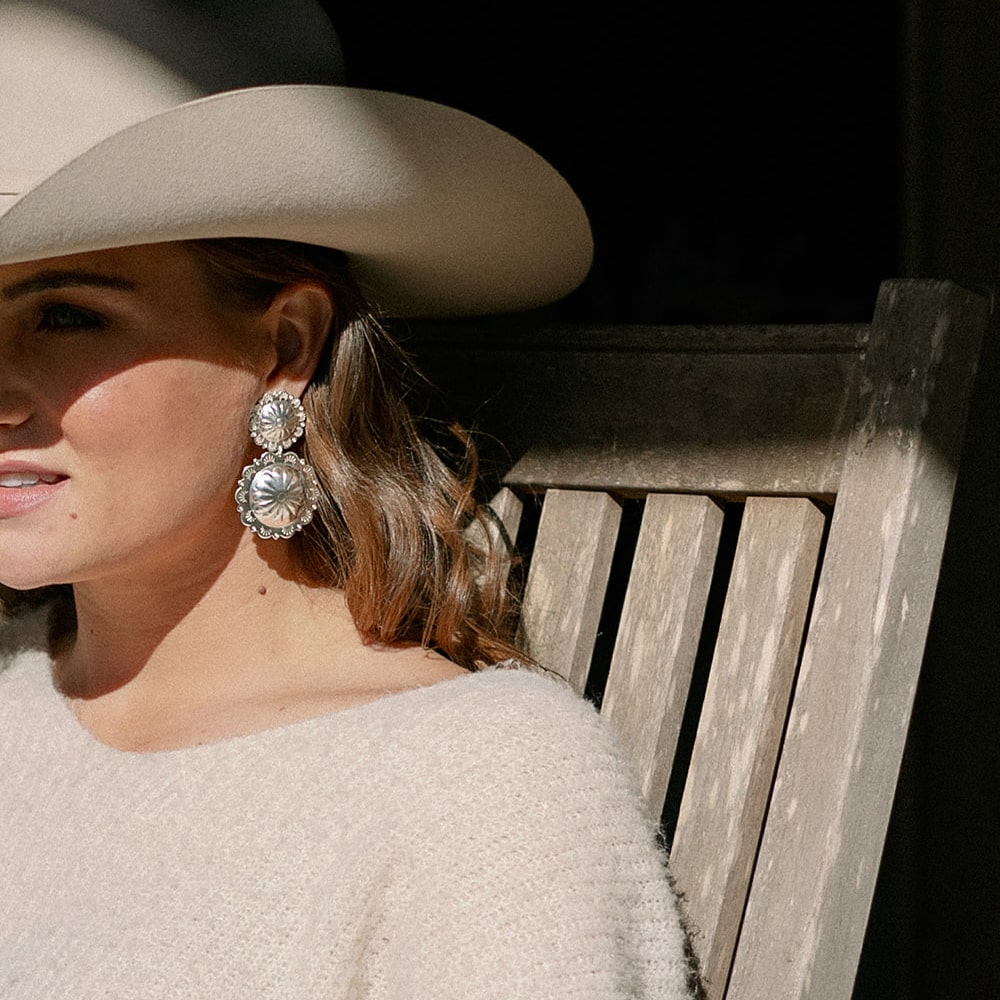 A woman wearing Concho Queen Dangle Earrings by Shop Marian sitting on a bench.