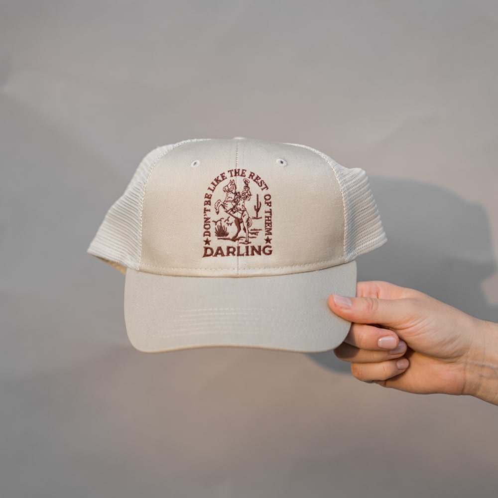 A hand holding a Darling Cap with the brand name Shop Marian, featuring an image of a tree.