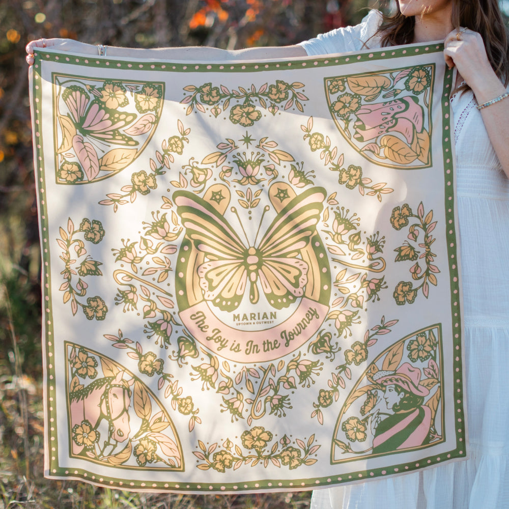 A woman triumphantly holds a Butterfly Scarf adorned with vibrant butterflies, symbolizing strength and growth amidst struggle, from Shop Marian.