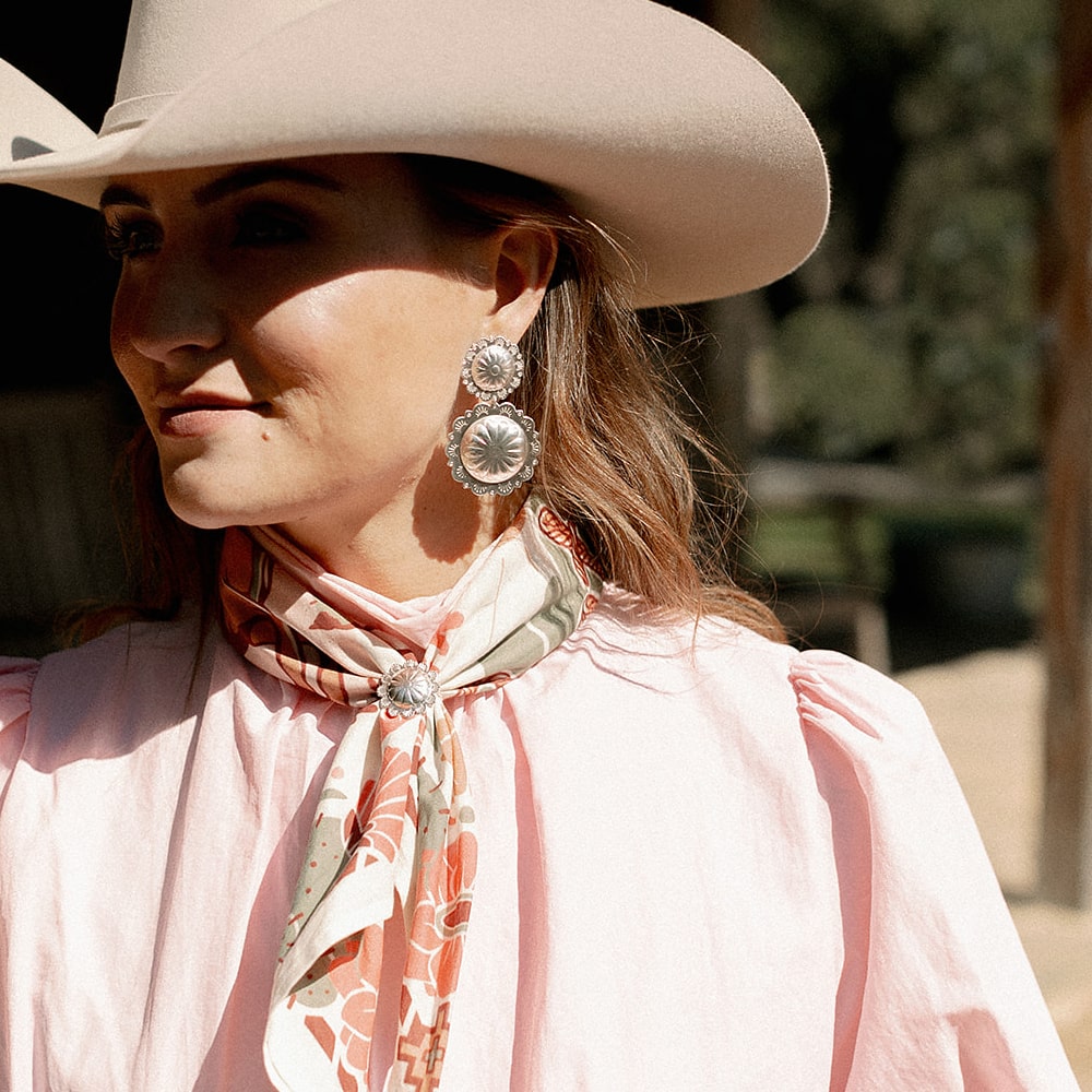 A woman wearing Shop Marian's Concho Queen Dangle Earrings and a cowboy hat and pink shirt.