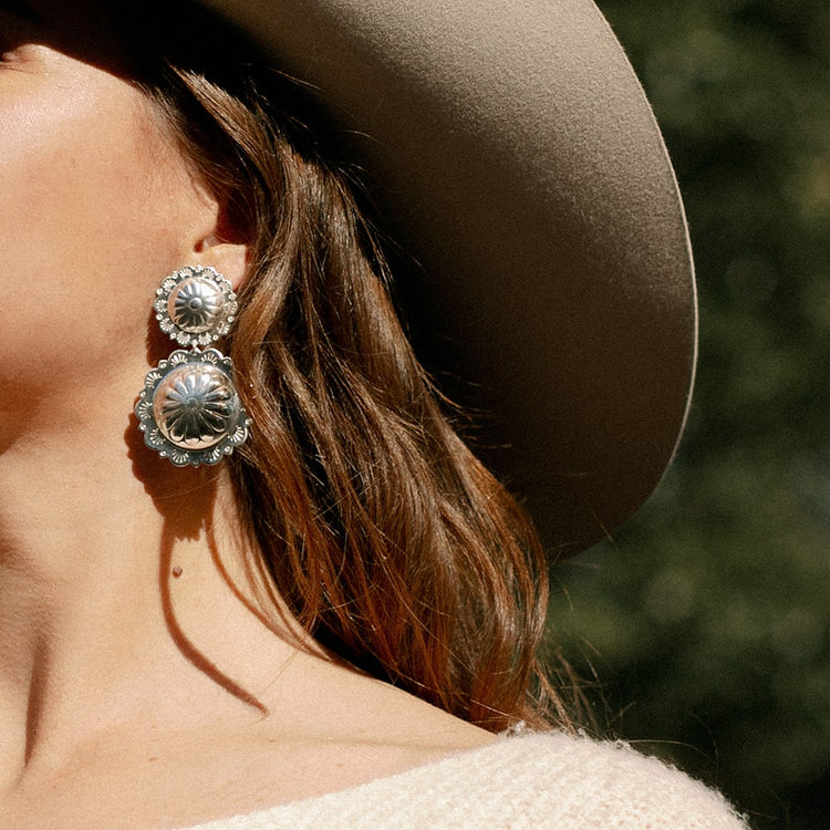A woman wearing a Concho Queen Dangle Earrings by Shop Marian and a hat.