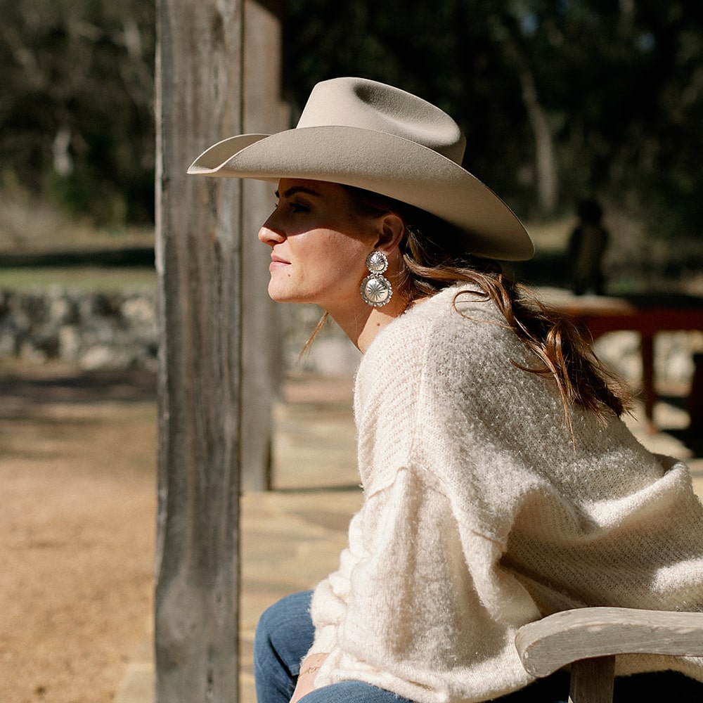 A woman in a cowboy hat sitting on a bench wearing Concho Queen Dangle Earrings from Shop Marian.