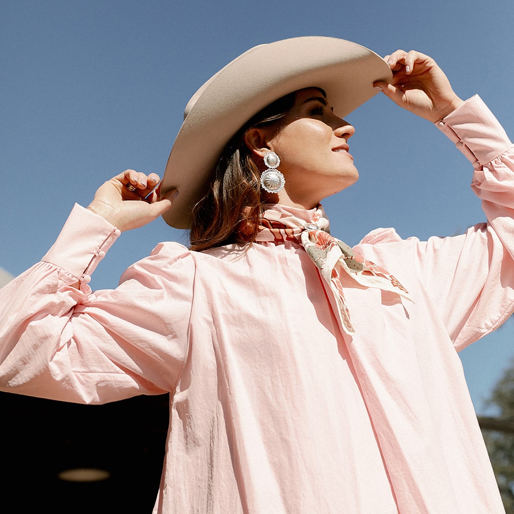A woman wearing Concho Queen Dangle Earrings by Shop Marian, a pink shirt and cowboy hat.