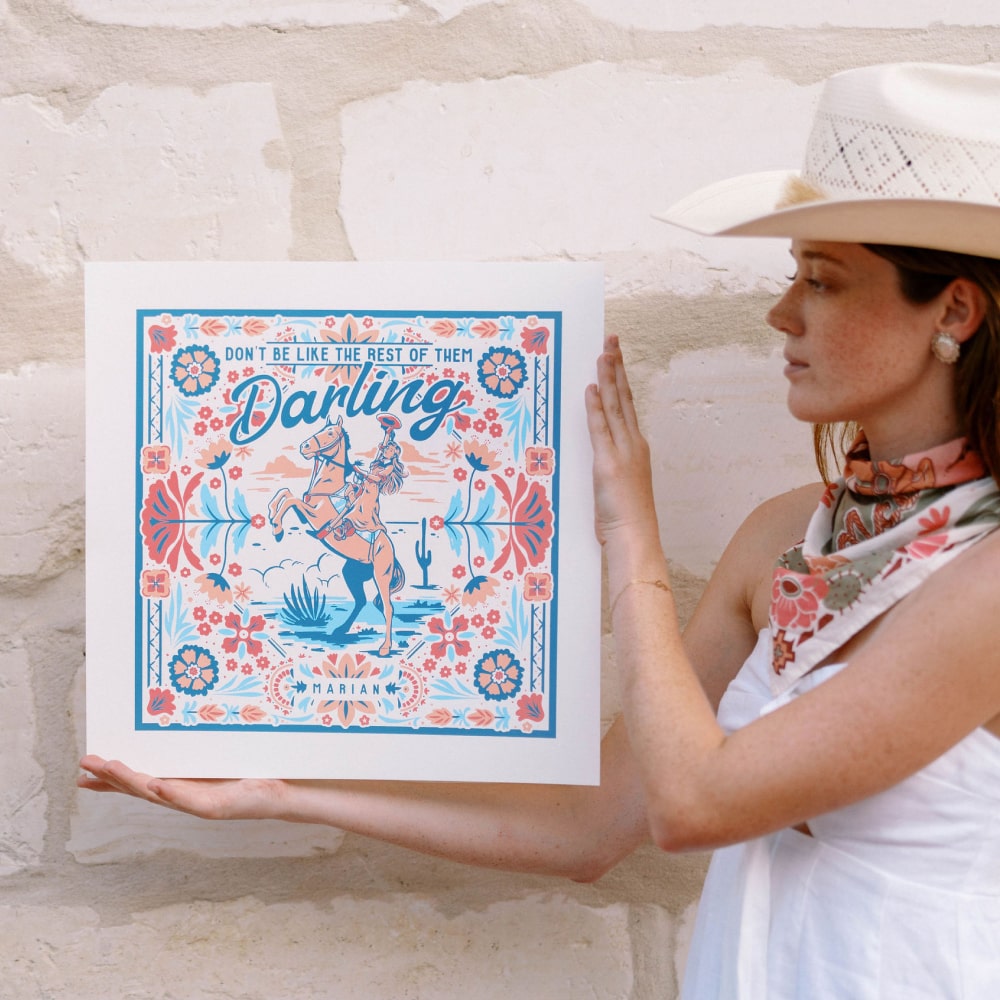 A woman in a cowboy hat holding up a Shop Marian Darling Print poster.