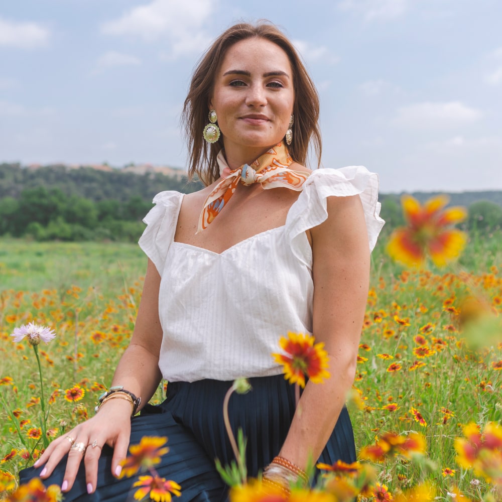 A woman sitting in a field of flowers wearing the Grow Good Things Scarf by Shop Marian.