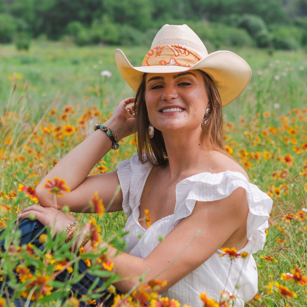A woman in a cowboy hat sitting in a field of flowers, wearing the Grow Good Things Scarf from Shop Marian.