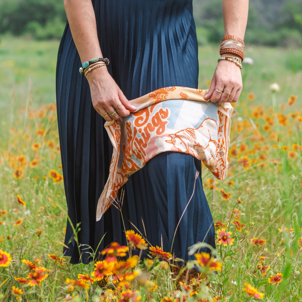 A woman holding a Grow Good Things Scarf by Shop Marian in a field of flowers.