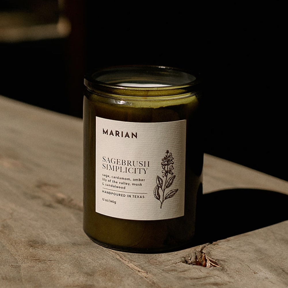 A Sagebrush Simplicity candle with a Shop Marian label on it sits on a wooden table.