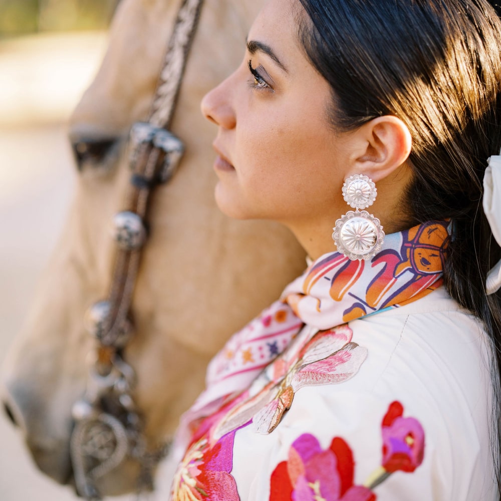 A woman wearing a ¡VIVA! scarf by Shop Marian and earrings next to a horse.