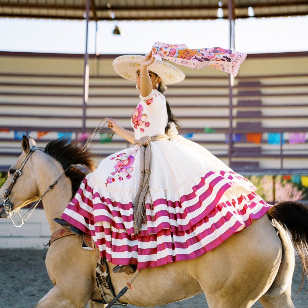 a woman in a Shop Marian mexican dress riding a horse wearing the ¡VIVA! Scarf.